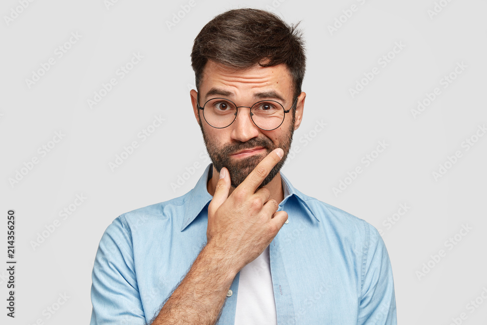 Image of hesitant unshaven European male with thick beard, holds chin, purses lips with clueless expressions, doubts what to eat delicious for supper, isolated over white background. What to choose? - obrazy, fototapety, plakaty 