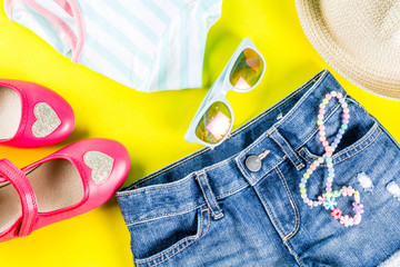 Summer vacation concept, summer child's cloth set - kids shorts, T-shirt, hat, sun glasses, bracelet necklace, sneakers, bright yellow background above flat lay copy space