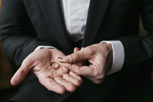 Wedding Rings On Palm Hand. Groom In Stylish Suit Holding Golden Wedding Rings In Hands, Sitting In The Room. Marriage Or Divorce Concept.