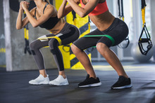 Close Up Of Athletic Women In Squat Together In Gym. Couple Of Fit Girls Are Exercising With Resistance Band For Lower Body Relief. They Are Wearing Sport Clothes And Sneakers