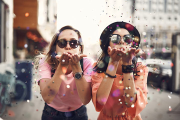 portrait of cheerful ladies blowing confetti from hands. they situating on street during sunny day. 