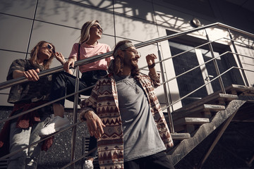 Wall Mural - Low angle of happy man with long hair locating near outgoing attractive young female on stairs. She leaning on handrail. Positive bearded dude situating opposite them