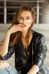 Lifestyle photo of young cheerful woman in fashion transparent glasses wearing a rock black style leather jacket touch her lips and look to camera. Hipster urban style. Lifestyle outdoor portrait.