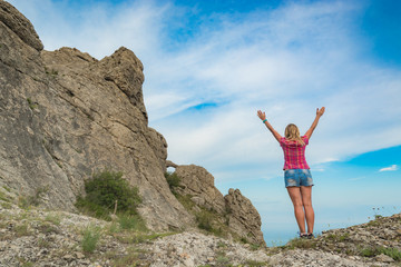 Wall Mural - Young happy woman stands with raised hands on a cliff