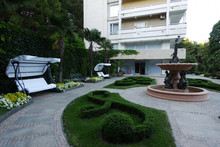 A Chic Interior Courtyard Near The Hotel With An Elegant Fountain With Sculptures By A Designer Lawn And White Benches With A Canopy With Beautiful Flowers Growing Next To It.