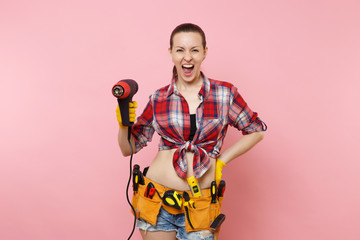 Young beautiful fun handyman woman in plaid shirt, denim shorts, kit tools belt full of variety instruments holding power heat gun isolated on pink background. Female in male work. Renovation concept.