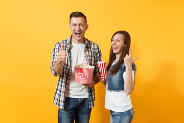  Young laughing couple woman man watching movie film on date hold bucket of popcorn plastic cup of soda cola showing thumb up pointing index finger up isolated on yellow background. Emotions in cinema.