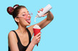 Fancy girl pressing whipped cream in red paper can. Posing with pipped tongue, smiling, wearing black tshirt, pink eyeglasses, pretty, cute hairstyle. Keeping white bottle. Blue studio background.