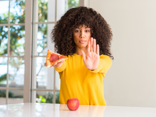 Poster - African american woman choosing between apple and pizza slice with open hand doing stop sign with serious and confident expression, defense gesture