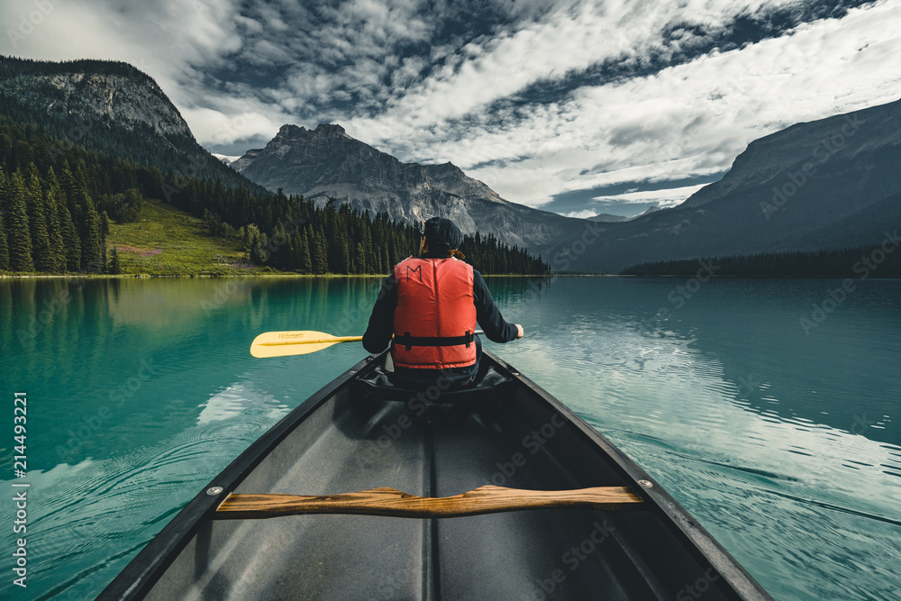 Young Man Canoeing on Emerald Lake in the rocky mountains canada with canoe and life vest with mountains in the background blue water. - obrazy, fototapety, plakaty 