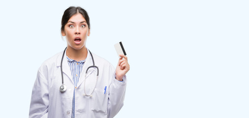 Sticker - Young hispanic doctor woman holding credit card scared in shock with a surprise face, afraid and excited with fear expression