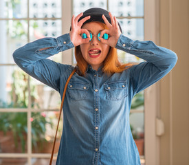 Sticker - Stylish redhead woman wearing bowler hat and sunglasses doing ok gesture like binoculars sticking tongue out, eyes looking through fingers. Crazy expression.