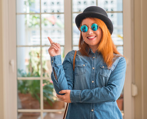 Sticker - Stylish redhead woman wearing bowler hat and sunglasses with a big smile on face, pointing with hand and finger to the side looking at the camera.