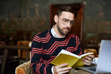 Fototapeta Dinusie - Young serious man in eyeglasses reading book while spending leisure in cafe