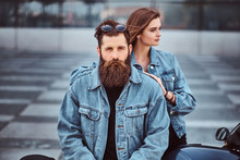 Close-up Portrait Of A Hipster Couple Of A Brutal Bearded Male And His Girlfriend Dressed In Jeans Jackets Against Skyscraper.
