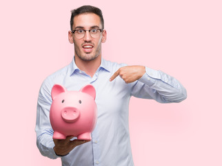 Canvas Print - Handsome young man holding a piggy bank with surprise face pointing finger to himself