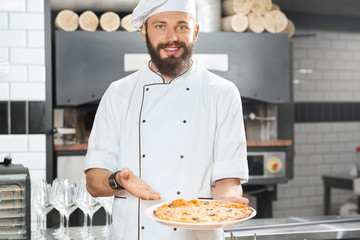 Smiling pizzaiolo holding fresh baked mouthwatering, delicious pizza. Wearing white chef's tunic working on restaurant kitchen with oven, wooden timbers, wine glasses set standing on background.