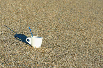 A cup of coffee in the sand at sunrise near the sea. Concept