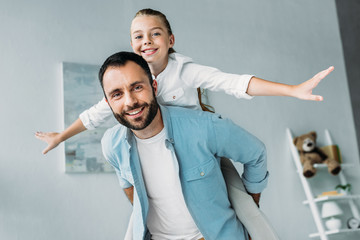 happy daughter piggybacking on father and looking at camera at home