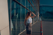 © WellStock - Beautiful blonde woman with curly hair, in stylish clothes looking on the sunset while standing near the big office center windows. Summer evening