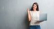 © Krakenimages.com - Young brunette woman over grunge grey wall using computer laptop surprised with an idea or question pointing finger with happy face, number one