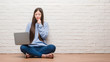 © Krakenimages.com - Young Chinese woman sitting on the floor over brick wall using laptop serious face thinking about question, very confused idea
