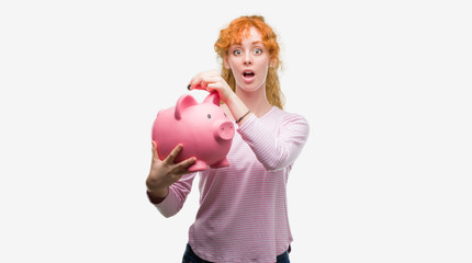 Canvas Print - Young redhead woman holding piggy bank scared in shock with a surprise face, afraid and excited with fear expression