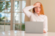 Redhead woman using computer laptop at home stressed with hand on head, shocked with shame and surprise face, angry and frustrated. Fear and upset for mistake.
