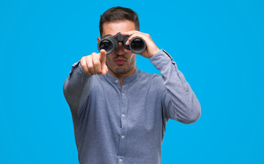 Sticker - Handsome young man looking through binoculars pointing with finger to the camera and to you, hand sign, positive and confident gesture from the front