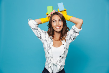 Sticker - Photo of optimistic smiling housewife 20s in yellow rubber gloves for hands protection holding two sponges during cleaning, isolated over blue background