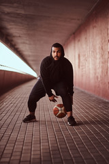 Wall Mural - Portrait of a dark-skinned guy dressed in a black hoodie and sports shorts playing basketball on a footway under bridge.