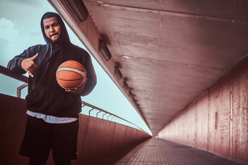 Wall Mural - Dark-skinned bearded guy dressed in a black hoodie and sports shorts leaning on a guardrail while standing with basketball.