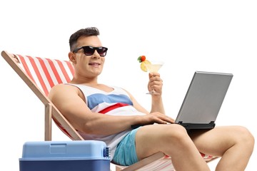 Wall Mural - Young man with a laptop and a cocktail sitting in a deck chair