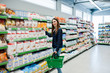 © AS Photo Family - Shopping woman looking at the shelves in the supermarket.  Portrait of a young girl in a market store holding green shop basket.