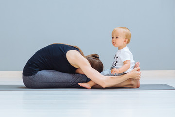 Wall Mural - Sportive brunette mother with baby son relax after good workout on grey yoga mat over gray wall background. Athletic and healthy motherhood. Fitness, happy maternity yoga with children concept.