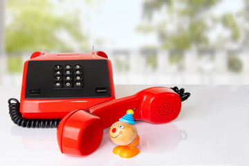 Red vintage telephone receiver with black cable on white table in front of natural blurred green background. Funny toy clown is taking a call. Suitable for contact exchange and call center. Template f