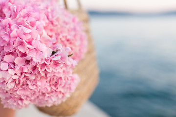 Wall Mural - Pink hydrangea are in straw travel bag. Girl holds flowers on background of sunset sea during vacation. Free space for text.