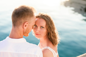 Couple of travelers are sitting by blue sea during romantic honeymoon vacation at sunset after wedding. Man and woman are happy tourists in resort. Lovers are wearing in white clothes outdoor.