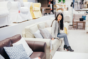 Beautiful girl choosing sofa in furniture store. Full length portrait of asian girl wearing jeans and gray coat sitting on light beige sofa