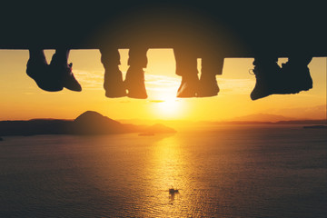 Wall Mural - Underside view of dangling Four people legs with wearing trekking shoes sitting on the edge of wooden board-walk against a background of sea and sunset.