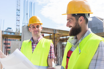 Wall Mural - Two young construction workers wearing yellow hard hats and reflective safety vests while analyzing together the plan of a new building