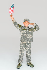 Wall Mural - little boy in camouflage clothing saluting while holding american flagpole in hand isolated on grey