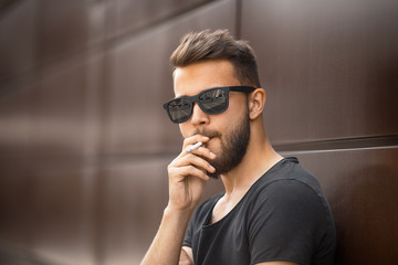 a young handsome white bearded man in sunglasses and black t-shirt smokes a cigarette in the street 