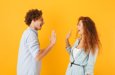 Photo of joyful couple man and woman standing face to face and giving high five, isolated over yellow background