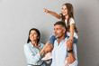 Photo of joyful family woman and man smiling and looking aside while pretty girl sitting on the neck of her happy father, isolated over gray background