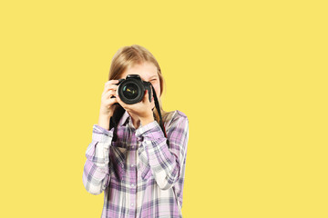 Young teenage girl holding digital photo camera with big lens & strap, taking pictures, smiling. Beautiful blond female photographer in checkered plaid shirt posing w/ dslr gear. Copy space background