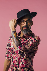 Vertical portrait of a very cool man with tattoos, tilting his hat, isolated on pink studio background