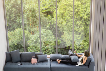 Asian woman laying on sofa near big glass wondows, relaxing alone in house with green forest in background