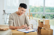 © DragonImages - Cheerful young ethnic man in casual clothes standing leaning on packed delivered cardboard box with clipboard and looking at mobile phone in hands smiling on blurred background of packed boxes in art