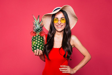 Poster - Happy young beautiful woman in red swimsuit, sunglasses and big summer hat with pineapple in sunglasses in hand posing over pink background